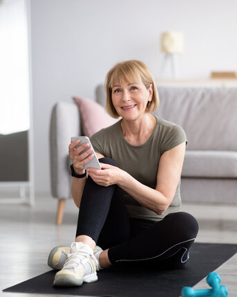Eine lächelnde Frau sitzt auf einer Trainingsmatte in einem modernen Raum. Sie trägt sportliche Kleidung und hält ein Smartphone in der Hand. Neben ihr befinden sich eine Wasserflasche und ein Paar Hanteln.