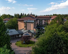 Ein mehrstöckiges Gebäude mit einem modernen Design, umgeben von Bäumen und Grünflächen. Das Gebäude hat ein schräges Dach und große Fenster, die viel Licht hereinlassen.