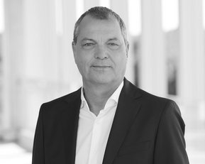 Mann in Anzug steht vor einer hellen, modernen Architektur. Er hat kurze Haare und schaut direkt in die Kamera.