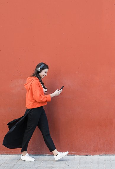 Frau läuft mit Handy vor roter Wand Eine Frau in einem orangefarbenen Hoodie und schwarzen Hosen geht an einer roten Wand entlang und schaut auf ihr Smartphone. Sie trägt Kopfhörer und weiße Schuhe.