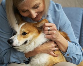 Eine Frau umarmt einen Corgi-Hund auf einem Sofa, lächelnd und liebevoll, während sie ihn streichelt.
