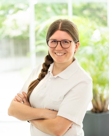 Eine lächelnde Frau mit langen, geflochtenen Haaren und Brille steht mit verschränkten Armen in einem hellen Raum mit Pflanzen im Hintergrund.