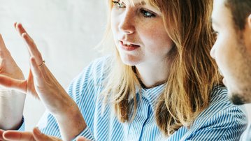 Eine Frau und ein Mann diskutieren an einem Computer, während die Frau mit ihren Händen Gesten macht. Beide scheinen an einem Projekt oder einer Präsentation zu arbeiten.