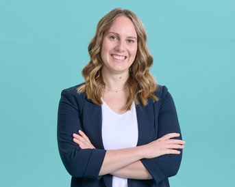 Eine lächelnde Frau mit lockigem Haar, die in einem blauen Blazer und einem weißen Oberteil gekleidet ist, steht mit verschränkten Armen vor einem türkisfarbenen Hintergrund.