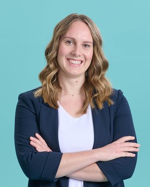 Eine lächelnde Frau mit lockigem Haar, die in einem blauen Blazer und einem weißen Oberteil gekleidet ist, steht mit verschränkten Armen vor einem türkisfarbenen Hintergrund.