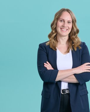 Eine lächelnde Frau mit lockigem Haar, die in einem blauen Blazer und einem weißen Oberteil gekleidet ist, steht mit verschränkten Armen vor einem türkisfarbenen Hintergrund.